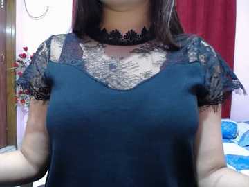 Video of a woman with light brown skin, wearing a black lace-trimmed blouse over a teal satin dress, standing indoors with a white wall, red curtain, and floral arrangement in the background.