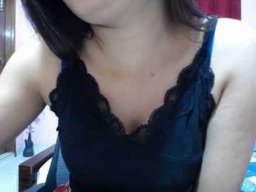 Video of a woman in a black lace-trimmed tank top, with shoulder-length dark hair. Background shows a wooden chair and a red curtain.