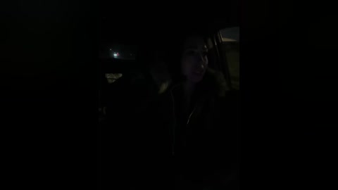A dimly lit video captures a young woman with long, dark hair and pale skin in a dimly lit car, wearing a dark coat, with a dim streetlight visible through the window.