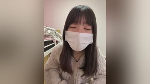 Video of an Asian woman with long black hair and bangs, wearing a white mask, beige sweater, and sitting on a bed with white metal frame in a dimly lit room.