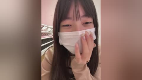 Video of an Asian woman with long black hair and bangs wearing a white surgical mask, standing in a bathroom with beige walls and a white metal shelf.