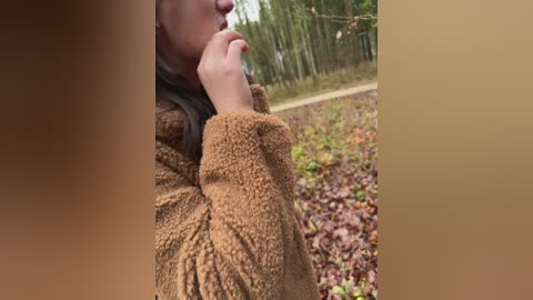 A close-up video of a person's right arm in a brown, textured, knitted sweater, with a hand resting on their chin. Background features a blurred, natural outdoor scene with greenery and a wooden fence.
