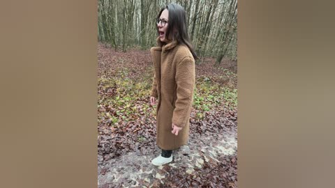 Video of a woman with long brown hair and glasses, wearing a brown coat and white sneakers, standing on a forest floor covered in fallen leaves.