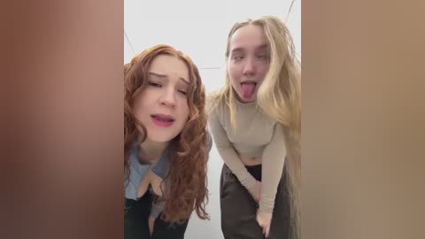 Video of two young women with long, curly red hair and blonde hair, respectively, in a narrow corridor. Both are leaning forward with playful expressions, tongue out.