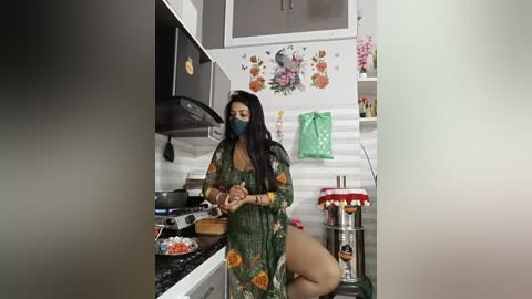 Video of an Asian woman with long black hair, wearing a green floral robe, cooking in a modern kitchen with floral wallpaper and stainless steel appliances.
