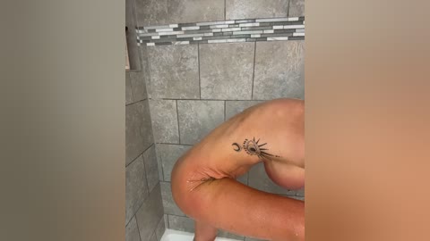 Video of a nude woman with fair skin and medium-sized breasts, bent over in a tiled shower with a mosaic accent strip. Her left thigh features a tattoo of a crescent moon and star.