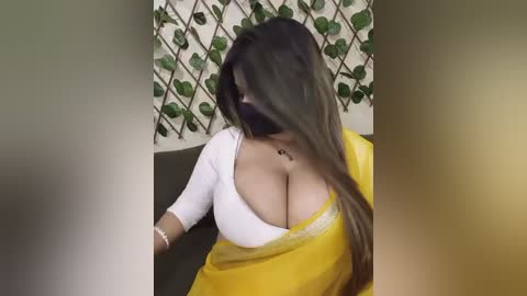 A video of a curvaceous woman with long dark hair, wearing a low-cut yellow sari and white top, partially obscuring her face. The background features a lattice-patterned wall.