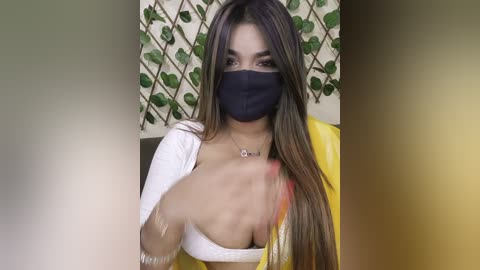 A video of a woman with long, straight brown hair, wearing a black mask, white top, and yellow sari, standing in a room with a green leaf patterned wall.