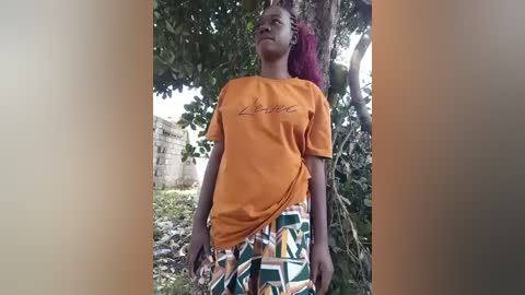 Video of a Black woman with pink braided hair, wearing an orange T-shirt with \"Liberia\" and patterned pants, standing outdoors among green foliage and a partially visible abandoned building.