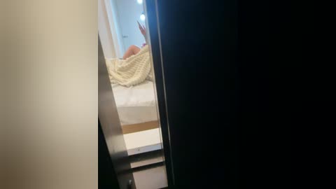 Media: Video of a person in a bed, covered with a light blanket, partially visible through a mirror, with bright lights reflecting in the background.