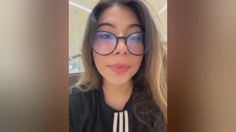 Video of a young woman with long, light brown hair and large, round glasses, wearing a black shirt with white stripes. Background features a beige wall and a blurred kitchen setting.