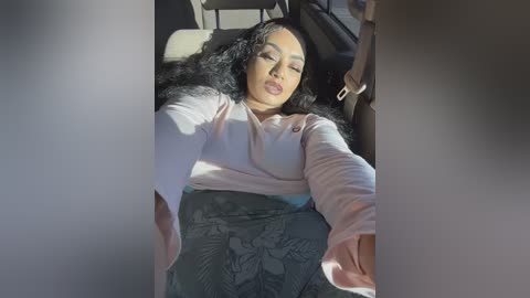 Video of a Black woman with long, curly black hair, wearing a light pink long-sleeve top and patterned gray skirt, asleep in a car seat, sunlight streaming in.