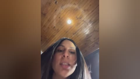 Video of a young woman with medium brown skin, long dark hair, and a warm expression, looking up at a wooden ceiling with a bright light bulb.