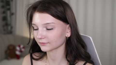 Video of a young Caucasian woman with long, dark brown hair, wearing minimal makeup, a black top, and small earrings. She has fair skin and a serene expression, seated indoors with a blurred background.