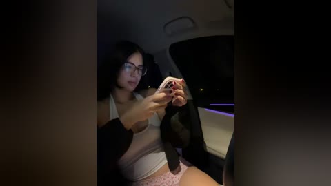 Video of a woman with dark hair, glasses, wearing a white tank top and pink panties, adjusting a seatbelt in a dimly lit car.