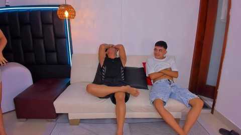 Video of two young men, one shirtless in a black tank top, the other in a white t-shirt and shorts, sitting on a white couch in a modern, dimly-lit room with a tufted headboard and a hanging lamp.