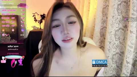 Video of a young, fair-skinned woman with long brown hair, smiling softly, taken in a dimly lit room with cream-colored curtains and a TV screen displaying a live chat.