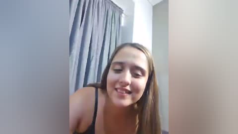 A video of a young woman with long brown hair, wearing a black tank top, leaning forward with a playful expression. She stands in a dimly lit room with gray curtains and beige walls.