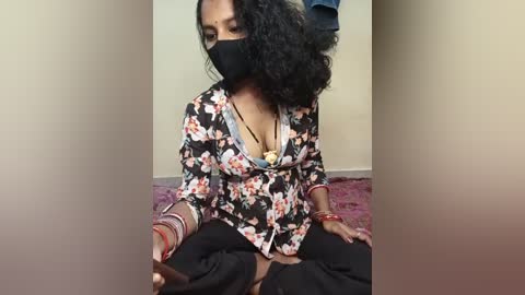 A video shows a South Asian woman with medium brown skin and curly black hair, wearing a floral blouse, black mask, and gold jewelry, sitting on a pink rug in a simple room.