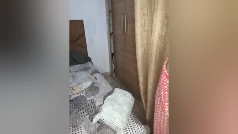 A video of a cluttered bedroom with a messy bed covered in various blankets and pillows. Wooden wardrobe with beige curtain on the right, white walls, and a partially visible person in the foreground.