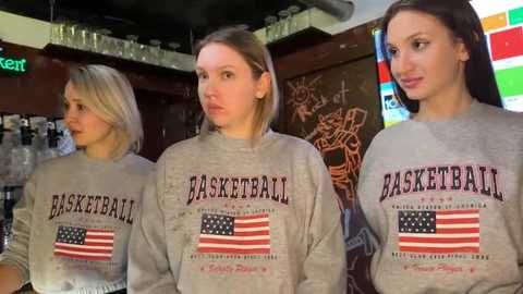 Video of three women wearing gray basketball jerseys with \"BASKETBALL\" in bold letters and an American flag design, in a dimly lit bar with glass shelves and colorful signage.