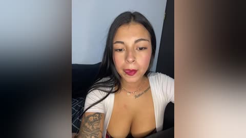 A video of a young woman with long black hair, wearing a white top exposing her cleavage, sitting on a dark couch, with blurred faces in the foreground.