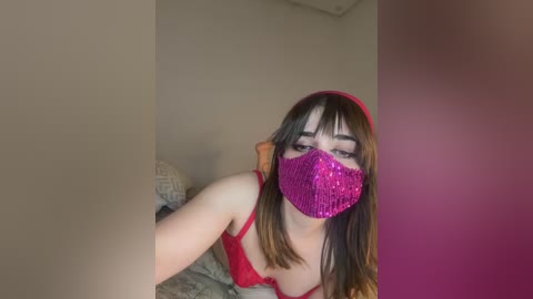 Video of a young woman with fair skin, straight brown hair, and a red headband. She wears a red lace bra and a pink glitter face mask, lying on a bed with beige walls and a gray quilt.