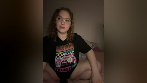A video of a young woman with curly hair, wearing a black \"Dazed and Confused\" t-shirt, sitting cross-legged on a bed, surrounded by dimly lit walls.