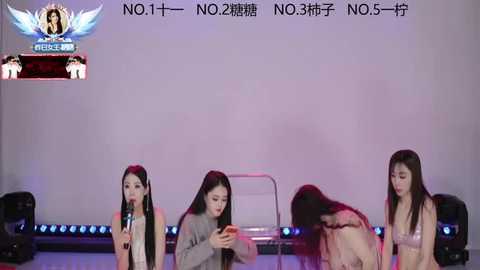 Video of four Asian women, long hair, singing on stage. Background has white text and promotional banners. Stage setup includes black chairs and mic stands.