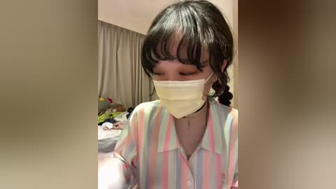 Video of a young Asian woman with dark hair in braids, wearing a pastel-striped blouse, face mask, and a black choker, standing indoors, blurred background.