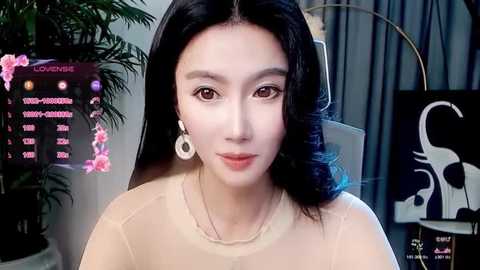 Media: Video of an East Asian woman with long black hair, wearing a sheer beige top and white earrings, standing in a modern room with digital screens and a potted plant.