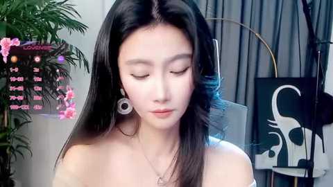 Video of an Asian woman with long black hair, wearing large circular earrings, standing in a modern room with a plant, clock, and abstract artwork.