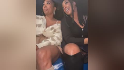 Video of two women seated closely, one in a white floral blouse and the other in a black jacket. They are smiling and appear to be engaged in conversation.