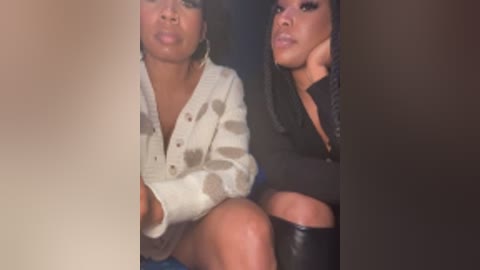 Video of two women with medium brown skin and dark hair, wearing white and black outfits, sitting closely, lips puckered, possibly in a dimly lit room.