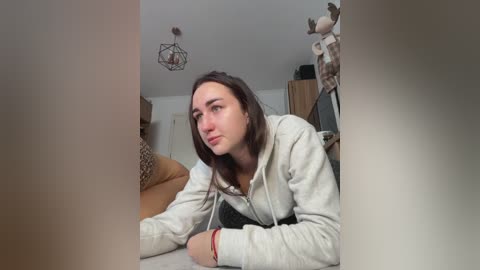 A video shows a young woman with fair skin, straight brown hair, and a neutral expression, lying on a couch in a cozy room with a wooden table and hanging lights.