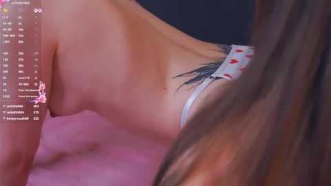 Video of a woman's bare back, with a visible tattoo of a black floral design on her lower back. She is wearing white panties with red hearts. The background is blurred, suggesting an indoor setting.