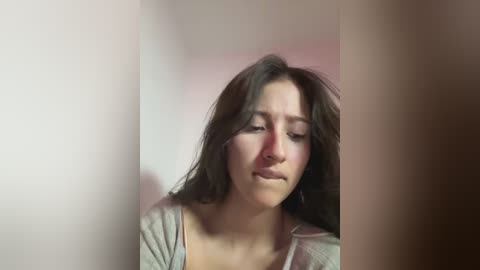 Media: Video of a young woman with long, dark hair, wearing a light gray shirt, looking down with a sad expression in a dimly lit room.