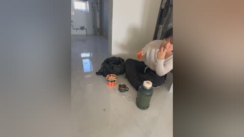 Video of a person sitting on a shiny white floor, wearing a grey sweater and black pants, eating food from a red bowl, surrounded by a black bag, orange bowl, and green bottle, in a dimly lit, bare room.