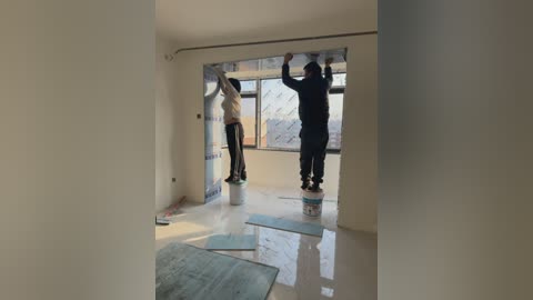 Video of two men in hard hats and protective gear, standing on plastic sheets, installing insulation in a bare room with large windows.