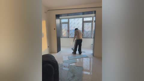 Video of a large, empty room with a man in a beige sweater and black pants cleaning the shiny white floor. The room has large windows with a cityscape view, and a black couch partially visible.