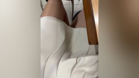 Video of a woman's legs clad in white leather boots and white leather skirt, seated on a wooden chair, viewed from a low angle, reflecting on a mirrored surface.