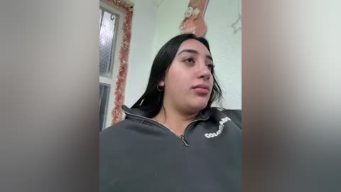 Video of a young Latina woman with long black hair, wearing a black hooded sweatshirt, standing indoors in a dimly lit room with peeling red and white walls and a window.