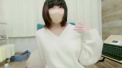 Video of a young Asian woman with shoulder-length black hair and a face mask, wearing a white sweater, seated at a desk in an office setting.