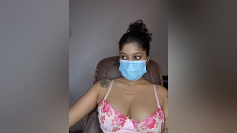 Video of a light-skinned woman with dark hair in a bun, wearing a blue face mask and a pink floral bra, sitting on a brown leather chair.