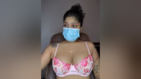Video of a young South Asian woman with medium-brown skin, wearing a blue mask, floral bra, and tattoos. She sits in a brown chair with a blurred background.