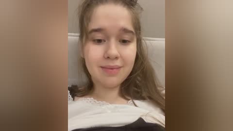 Video of a young girl with light skin, long brown hair, and a white and black dress, smiling slightly, against a plain beige background.