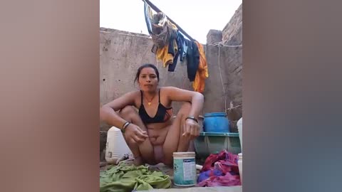 Video of a woman in a small, dirty courtyard with clothes hung on a line, wearing a black bikini, squatting, and surrounded by laundry, buckets, and colorful fabric.