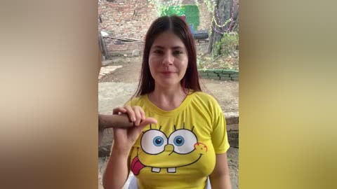 Video of a woman with long brown hair, wearing a bright yellow SpongeBob T-shirt, holding a man's hand, in a garden with greenery and a brick wall background.