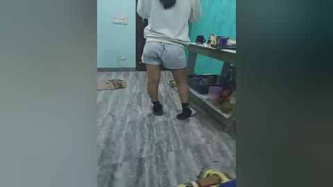 A video of a person from behind, standing in a messy room with light wood flooring. They wear gray shorts and a white sweatshirt, black socks, and black sneakers.