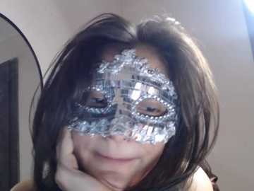 Video of a woman with long dark hair, wearing a silver, ornate masquerade mask with rhinestones, smiling slightly, with a blurred background of a room with a round mirror.